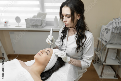 Aesthetician uses a dermapen device to provide a microneedling session for a young woman seeking glowing, healthy skin. The clean, bright clinic environment highlights the focus on beauty and wellness