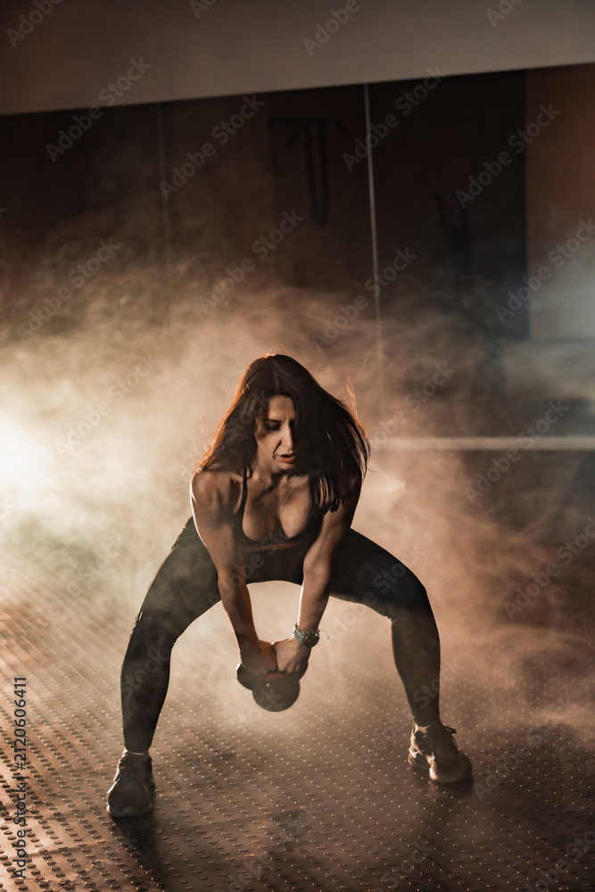 Fototapeta premium Determined woman in sportswear trains with a kettlebell, surrounded by dramatic smoke and gym lighting. Fitness motivation and strength training concept for modern active lifestyles