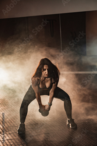 Determined woman in sportswear trains with a kettlebell, surrounded by dramatic smoke and gym lighting. Fitness motivation and strength training concept for modern active lifestyles
