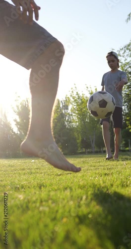 Barefoot boy passes football with father in park at sunset. Child practices soccer skills on grass while only legs of parent are visible in vertical video. Father and son playing football.