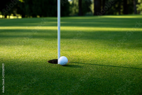 White golf ball tap in close to hole and pin on putting green, early morning sunny summer day on the golf course, relaxing recreational sport

