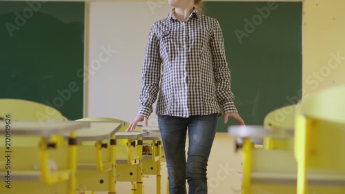 A pregnant woman is seen standing in a bright, empty classroom, surrounded by yellow desks and a green chalkboard. The scene highlights maternity in a contemporary educational setting