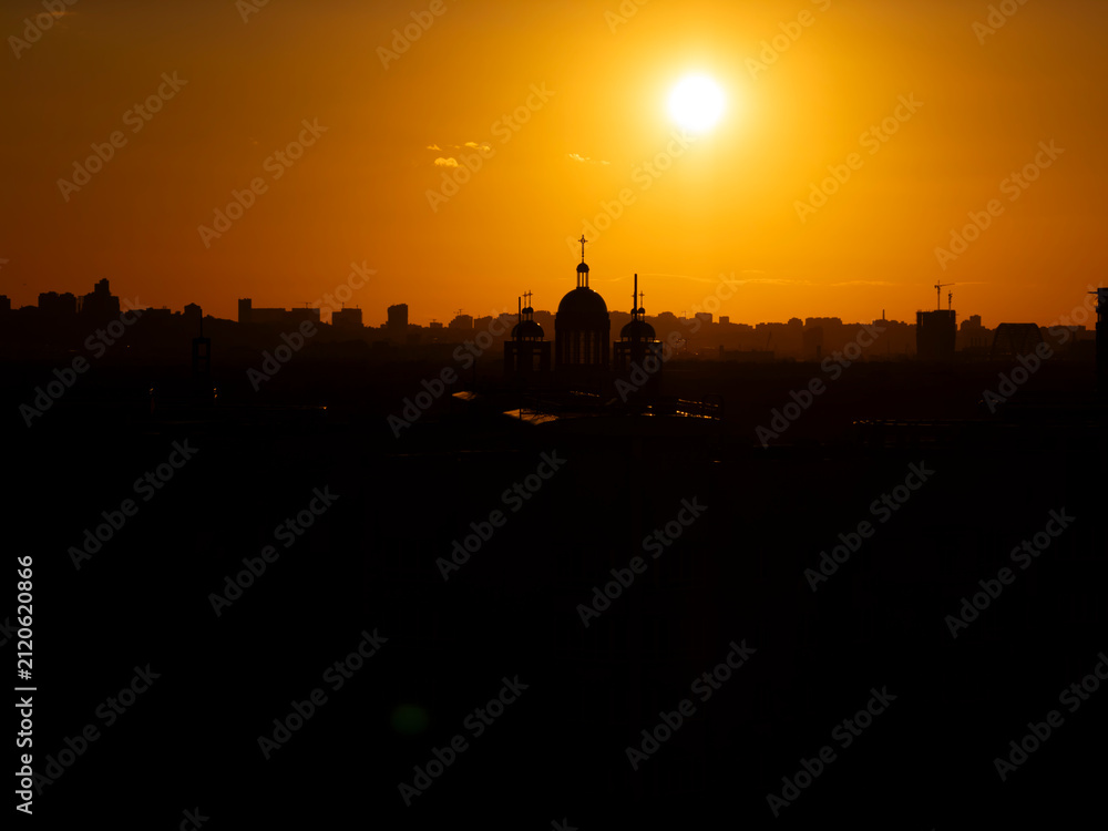 Obraz premium Urban cityscape showing building silhouettes and church domes against a bright orange sky during a vibrant sunset, representing tranquility and the beauty of evening light over a developing city