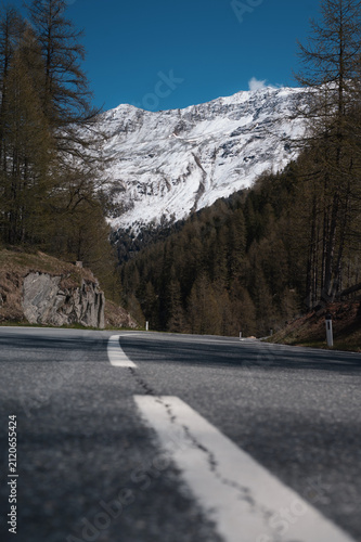 Low perspective of a mountain road with center line leading toward snowy alpine peaks and forest in Tyrol, Austria.