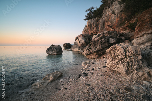 Calm sunrise seascape with rocky beach, limestone cliffs, clear water, and coastal scenery at Cala Fuili in Sardinia.