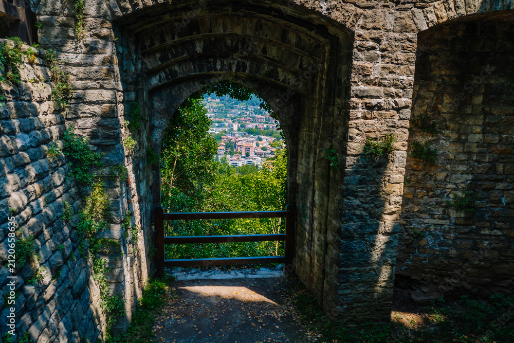 Obraz premium Stone archway leading to a scenic view of Bergamo city from Rocca di Bergamo park, surrounded by greenery and historic architecture