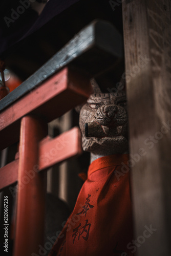 A weathered stone fox statue wearing a red bib with Japanese characters, partially lit beside a red torii gate structure at an Inari shrine.