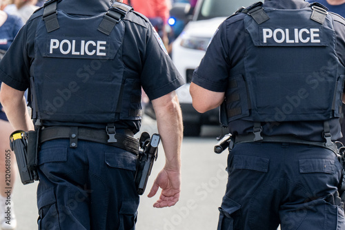 Two police officers wearing blue uniforms with white lettering on their backs. The cops are wearing guns, handcuffs, tasers, protective bulletproof vests, tactical belts, and narcotic testing kits.