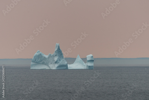 A large white iceberg formation floating in the cold ocean with layers of textured ice and snow. The ice is in transition, melting from the warm rays of the sun at sunset. The berg is a blocky shape.