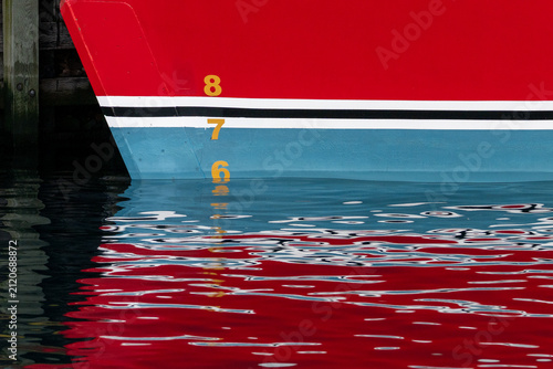 A red ship with waterline marks painted white, blue and black on the bottom of the boat. The gold numbers are painted on the bow of the boat. The numbers and lines are reflected in the calm water.
