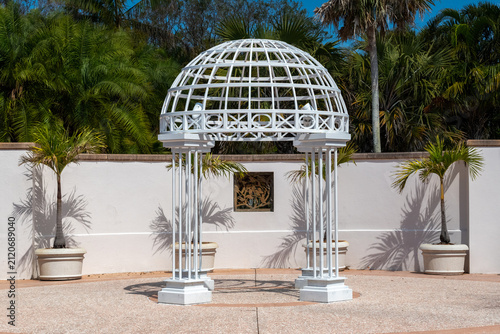 A large white metal gothic-style archway dome in an enclosed outdoor garden. The wedding structure has four heavy-duty iron pillars. The background is lush green trees and shrubs. The ground is a bric