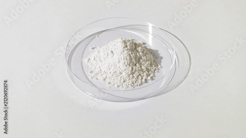 Vertical jib shot of white fine powder piled in a plastic petri dish on a pale gray canvas lit by moving light creating dynamic shadows | Titanium dioxide cosmetic ingredient concept