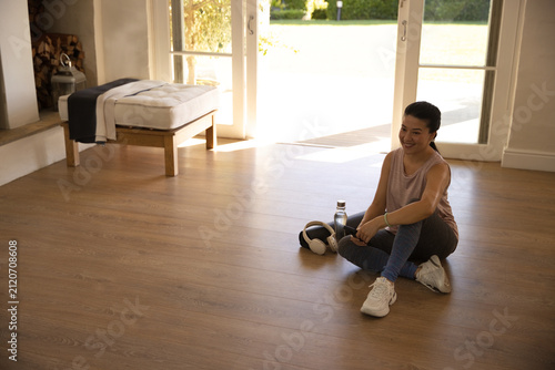 Asian woman sitting on wood floor by doors in sneakers  water bottle  headphones  copy space