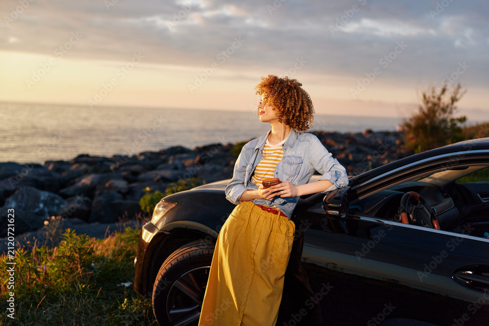 Fototapeta premium A woman in a denim jacket and mustard skirt leans against a sleek car by the coast at sunset, embracing the breeze. A candid lifestyle moment of outdoor freedom.