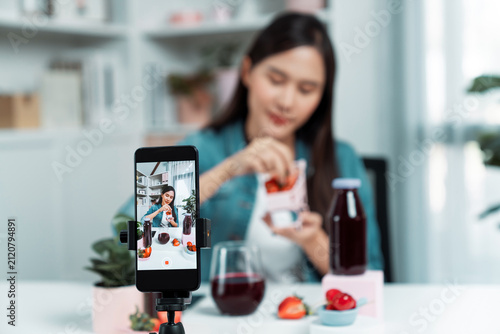 Foto Selective focus smartphone with smiling young beautiful Asian reviewing mixed strawberry and cherry juice detox drink, presenting healthy product promotion selling on social media online