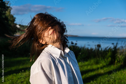 Foto Young woman in a white shirt with windblown hair posing near a lake on a sunny summer day