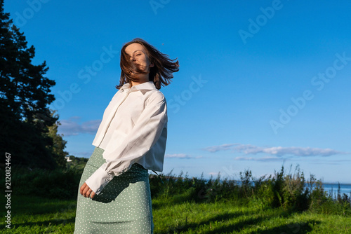 Bild auf Leinwand Young woman in a white shirt and green polka dot skirt with windblown hair posing near a lake on a sunny day