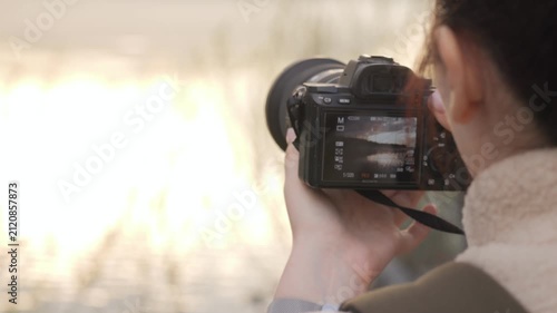 Person holds camera, focusing on bright outdoor scene. Back view shows intent to capture natural light. Soft focus background suggests serene environment. Ideal for travel, photography