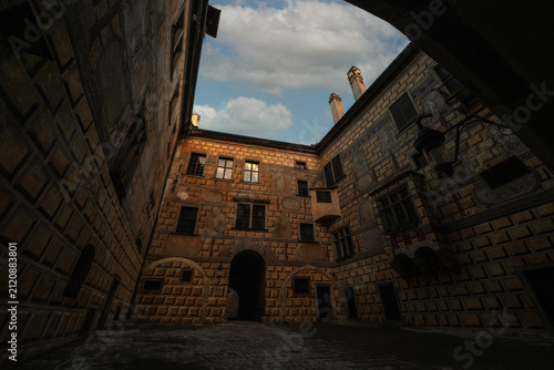 dramatic low-angle view of a historic castle courtyard featuring ornate Renaissance sgraffito facades, warm sunlight, and an arched gateway under a cloudy blue sky.