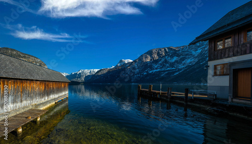 A serene view of Lake Hallstatt in Austria, framed by traditional wooden boathouses and a dock, with majestic snow-capped mountains reflecting in the clear water under a brilliant blue sky.