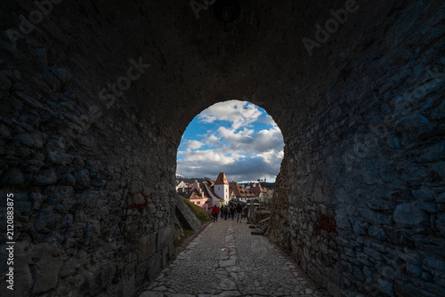 A dark historic stone archway framing a view of a charming old town street with traditional buildings, cobblestone paths, and people walking under a bright cloudy blue sky