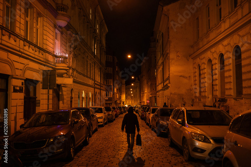 A lone man walking down a historic cobblestone street lined with parked cars and glowing old buildings at night urban atmosphere.