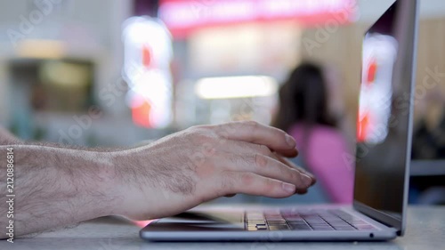 Hand types on laptop keyboard in public space. Background shows blurred cafe patrons and glowing signs. Focus stays on fingers and laptop. Person appears focused and relaxed while working