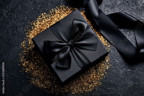 Black gift box with silk bow resting on gold glitter over dark textured stone in a luxurious macro shot for holiday and luxury retail marketing