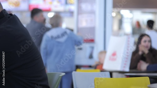 Blurry indoor scene with people sitting and standing near tables. One person points at a menu board. Others chat or wait. Yellow chairs add color to space. Suitable for cafe, restaurant