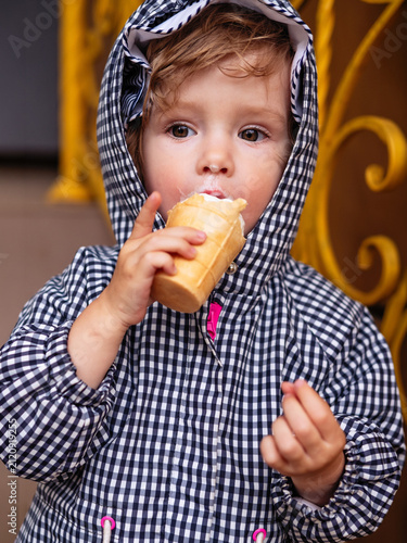 A toddler wearing a checkered hooded jacket enjoys a vanilla ice cream cone outdoors. The close-up portrait captures a curious child with a creamy ice cream mustache, creating a candid