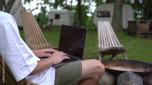 Person typing on laptop while seated in a hammock. Background shows campsite with trailers and firepit. Person appears relaxed, focused on work or leisure. Ideal for remote work, travel