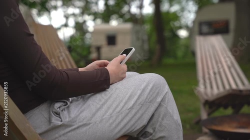 Person relaxes on wooden chair, using smartphone outdoors. They sit calmly, focused on phone screen. Background shows grassy yard and campfire pit. Ideal for scenes of leisure, digital connection
