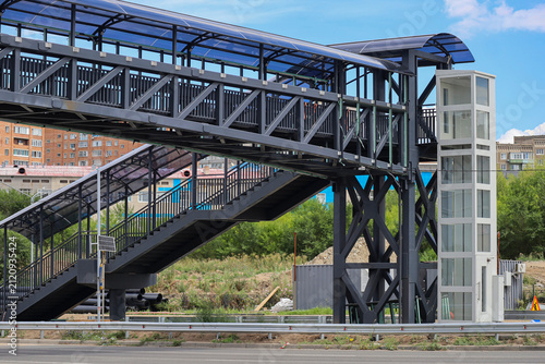 Modern covered pedestrian overpass with elevator, steel construction and transparent roof above a city roadway. Contemporary urban infrastructure.