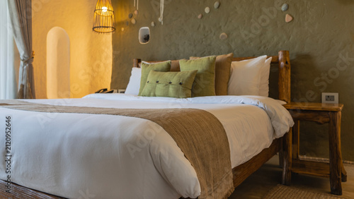 The bedroom interior is rustic. The bed is made up. Pillows at the headboard. Log bedside tables. The wall is roughly finished and decorated with pebbles. The lamp is lit.