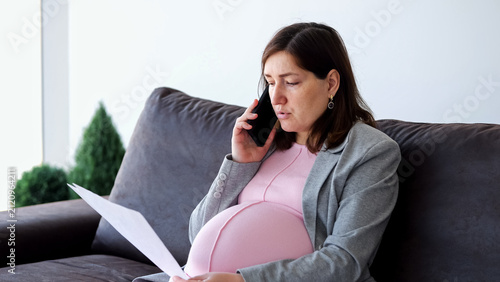 Pregnant businesswoman wearing a blazer and pink maternity dress, sitting on a sofa at home, talking on a smartphone while holding and reviewing documents