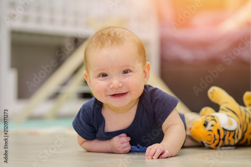 Smiling baby boy wearing a blue romper crawling on a wooden floor, looking directly at the camera with a cheerful expression next to a plush tiger toy in a bright room