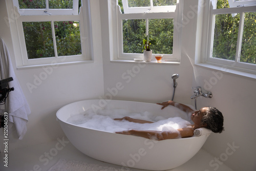 Freestanding oval bathtub is filling with foamy bubbles in bright bathroom near windows