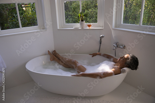 Oval freestanding bathtub is holding bubbly water in bathroom corner near bay windows and plant