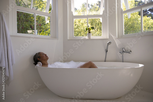 Freestanding oval tub is sitting beneath three sash windows, bubbles rising beside chrome faucet
