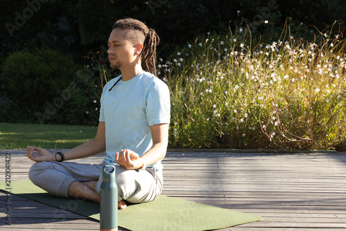 Fotografía African American man meditating on sage-green yoga mat on wooden deck with water