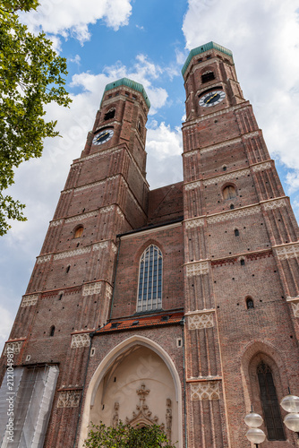 Fotografía The twin brick towers with green copper domes and clock faces of the Frauenkirche in Munich, Germany, seen from below against a summer sky