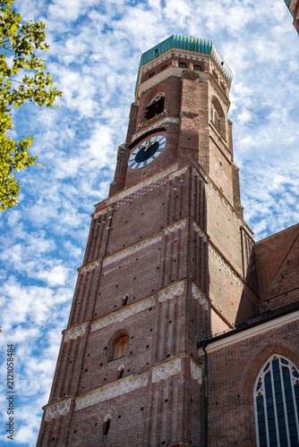 Fotografía Single brick tower of a Gothic cathedral with a green copper dome and black clock face rising into a bright cloud filled sky