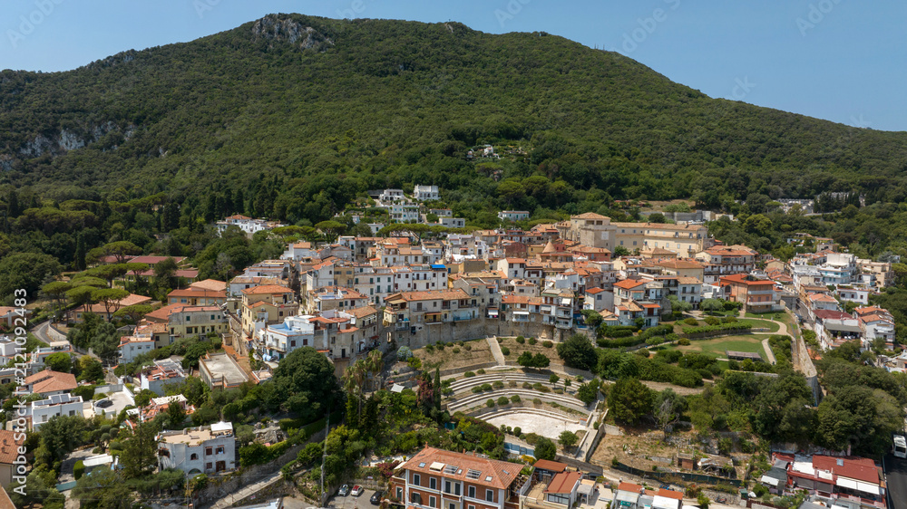 Fototapeta premium Scenic aerial view of San Felice Circeo historic town, showing charming houses with red roofs nestled on a green hillside with an amphitheater, beneath a lush mountain under a bright summer sky.