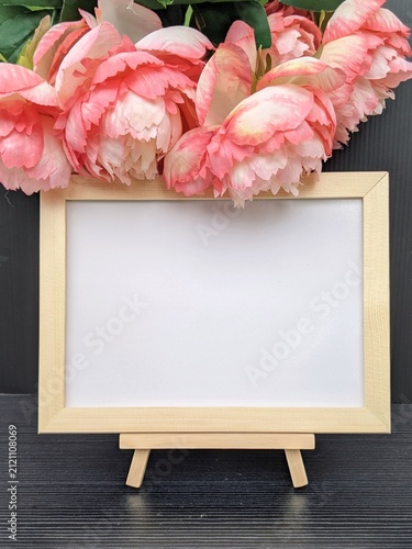 Charming wooden frame on an easel surrounded by vibrant pink peonies, creating a beautiful and artistic display, perfect for showcasing messages, photographs, or art pieces in a cozy, elegant set