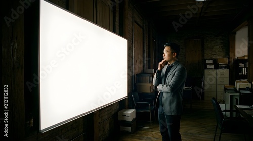 Asian man standing in dark office looking at glowing blank white screen