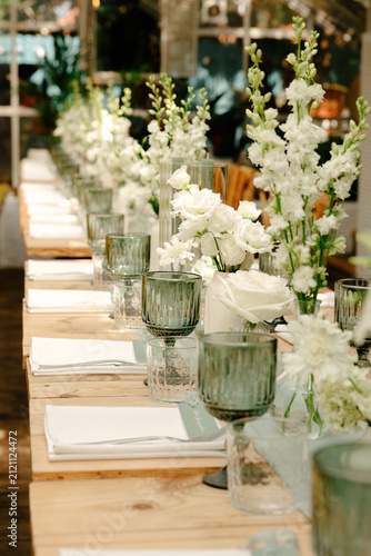 Wedding table running toward bright greenhouse windows with a row of tall white blooms in glass vases, greenhouse venue marketing, wedding florist portfolio, event planning website imagery, nobody