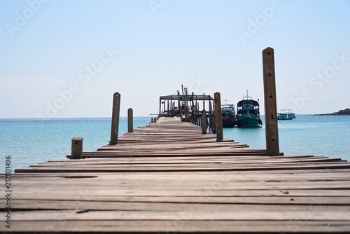 Wooden Jetty Extending into Turquoise Waters: A rustic wooden jetty juts out into the calm, clear turquoise sea, with boats moored at the end, under a vast, cloudless sky.