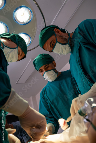 Operating room surgery team under bright lamp with masked surgeon in green scrub