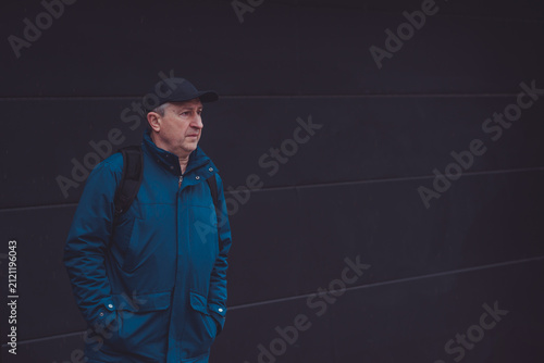 Thoughtful senior man aged 50s 60s in blue jacket and cap standing alone outdoors looking into distance, psychological mental health concept of loneliness and reflection