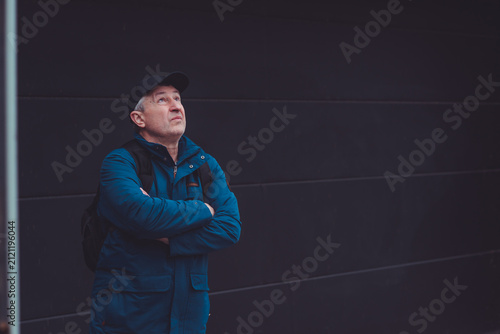 Thoughtful senior man aged 50s 60s in blue jacket and cap standing alone outdoors looking into distance, psychological mental health concept of loneliness and reflection